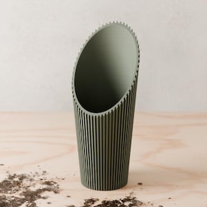 Peut inclure: Un pot de fleurs cylindrique vert sauge avec des rainures verticales et un bord supérieur incliné. Le pot est fait d'un matériau solide et repose sur une surface en bois clair. Le design est moderne et minimaliste.