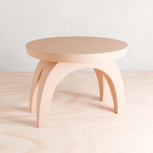 May include: A light pink round table with three arched legs. The table is made of wood and has a smooth surface.