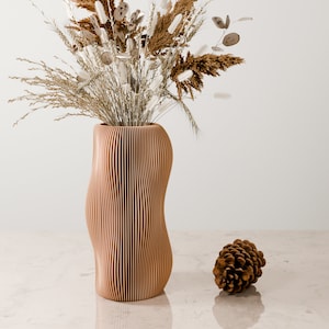 May include: A wooden vase with a wavy, ribbed design, holding dried flowers and grasses. The vase is light brown and has a natural wood finish. A small pine cone sits on the table in front of the vase.