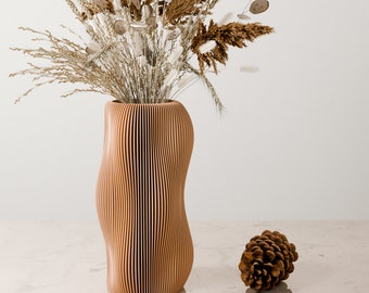 Puede incluir: Un jarrón de madera con un diseño ondulado y acanalado, que contiene flores y hierbas secas. El jarrón es de color marrón claro y tiene un acabado de madera natural. Un pequeño cono de pino se encuentra en la mesa frente al jarrón.