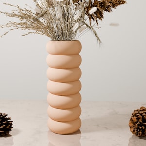 May include: A light peach-colored ceramic vase with a unique, rounded, stacked design. The vase is filled with dried grasses and flowers.