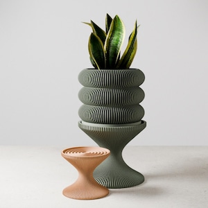 Puede incluir: Un macetero de cerámica verde con un diseño acanalado y un soporte a juego. El macetero está lleno de una planta verde con rayas amarillas. Un soporte de cerámica naranja más pequeño con un diseño acanalado similar está en primer plano.