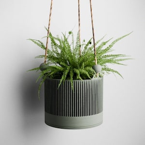 May include: A hanging planter with a green fern. The planter is cylindrical with vertical lines and a solid base. It is suspended by three brown cords with gray spherical accents. The fern has vibrant green fronds.