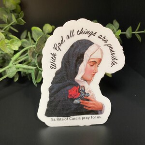 Puede incluir: Una pegatina blanca con una imagen de una mujer con hábito negro sosteniendo una rosa roja. El texto "With God all things are possible." está escrito en un círculo alrededor de la mujer. El texto "St. Rita of Cascia, pray for us." está escrito debajo de la mujer.