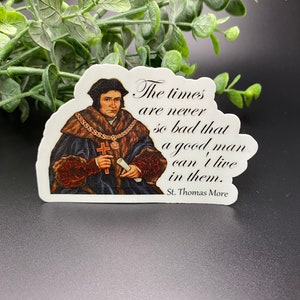 Puede incluir: Una pegatina con un retrato de Santo Tomás Moro, una figura histórica, con una cita: "Los tiempos nunca son tan malos que un buen hombre no pueda vivir en ellos."  Santo Tomás Moro.
