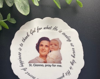 Puede incluir: Una pegatina blanca con una foto de una mujer sosteniendo un bebé. La pegatina tiene el texto: "El secreto de la felicidad es agradecer a Dios por lo que nos envía cada día en su bondad. Santa Gianna, reza por mí."