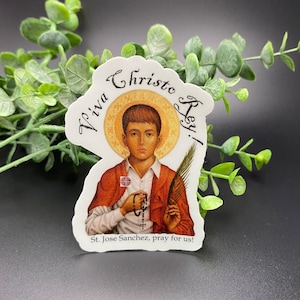 Puede incluir: Una pegatina con una imagen religiosa de un niño pequeño con cabello castaño y una túnica roja. El niño sostiene un rosario y una rama de palma. La pegatina tiene el texto "Viva Christo Rey!" y "St. Jose Sanchez, reza por nosotros!"
