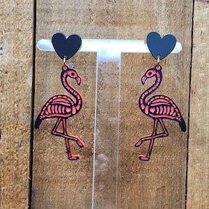 May include: A pair of black heart-shaped earrings with pink flamingo charms. The flamingos are skeletonized and have a black outline.
