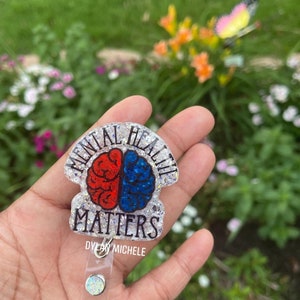 May include: A clear glitter badge holder with a silver glitter brain design. The brain is half red and half blue. The text "Mental Health Matters" is written around the brain. The badge holder has a clear retractable badge reel with a silver glitter button.