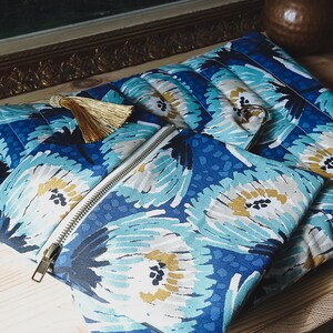 May include: A set of three blue and white floral patterned zippered pouches with gold tassels. The pouches are made of fabric and have a quilted texture.