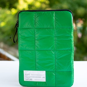Puede incluir: Funda acolchada para portátil de color verde con una etiqueta blanca que dice "SOFT IN NATURE".