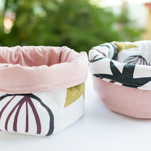 May include: Two fabric storage baskets with pink lining and white fabric with a geometric pattern in black, green, and purple.