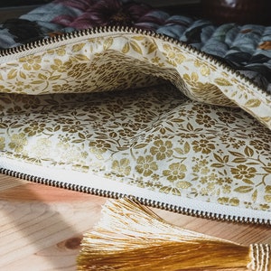 May include: A zippered pouch with a floral print on the outside and a gold floral print on the inside. The pouch is open and a gold tassel is hanging from the zipper pull.