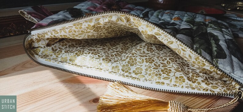 May include: A zippered pouch with a floral print on the outside and a gold floral print on the inside. The pouch is open and a gold tassel is hanging from the zipper pull.