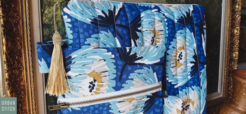 May include: A blue and white floral patterned zippered pouch with a gold tassel. The pouch is quilted and has a white zipper.