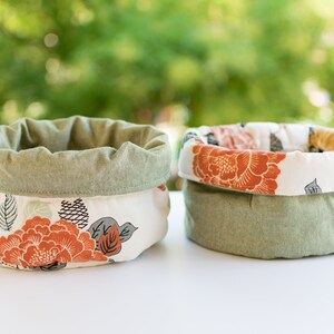 May include: Two fabric storage baskets with a floral print. The baskets are made of a green and white fabric with a floral pattern. The baskets are empty and sitting on a white surface.