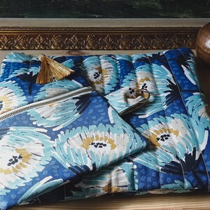 May include: Three blue and white floral patterned zippered pouches with gold tassels. The pouches are arranged on a wooden surface.