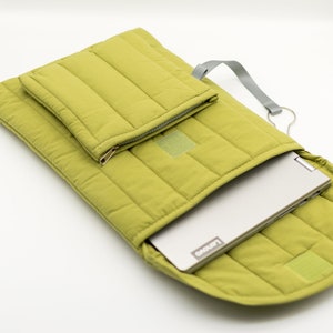 Könnte beinhalten: Eine grüne, gesteppte Laptoptasche mit einer Reißverschlusstasche auf der Vorderseite. Die Tasche ist offen und ein silberner Laptop befindet sich im Inneren.
