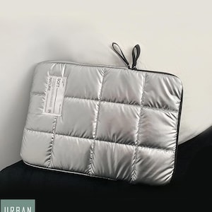 Op de afbeelding: Een zilveren, gewatteerde laptophuls met een zwarte rits en een klein wit label met de tekst "SOFT NATURE IN".