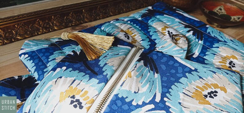 May include: A blue and white zippered pouch with a gold tassel. The pouch has a floral pattern with blue, white, and yellow flowers.
