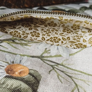 May include: A white canvas zippered pouch with a floral print featuring green leaves, white flowers, and orange centers. The pouch is lined with a white fabric with a small gold floral pattern.