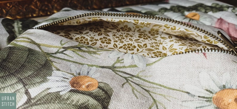 May include: A white canvas zippered pouch with a floral print featuring green leaves, white flowers, and orange centers. The pouch is lined with a white fabric with a small gold floral pattern.