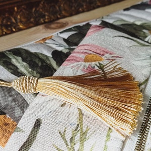 May include: A white canvas zippered pouch with a floral print featuring daisies and leaves. The pouch has a gold zipper and a gold tassel.