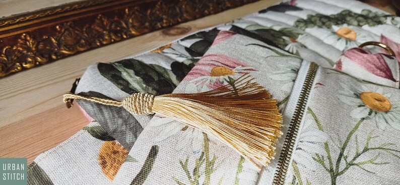 May include: A white canvas zippered pouch with a floral print featuring daisies and leaves. The pouch has a gold zipper and a gold tassel.