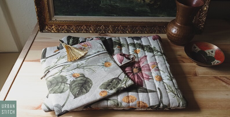 May include: Two floral patterned zippered pouches, one larger than the other, with a gold tassel on the smaller pouch. The pouches are made of a light beige linen fabric with a quilted design.