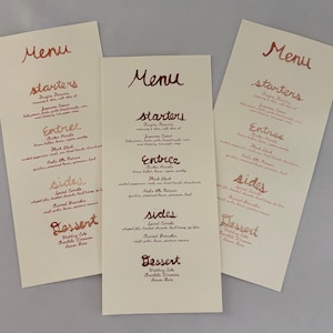 Op de afbeelding: Drie hoge, rechthoekige papieren menukaarten met een gebroken witte achtergrond en koperkleurige handgeschreven tekst. Elk menu bevat secties voor starters, hoofdgerechten, bijgerechten en desserts met gerechten zoals Foxfire Focaccia.