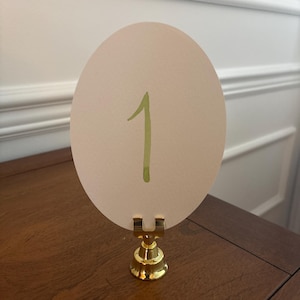 May include: A table number card with the number "1" in green watercolor on an oval, cream-colored card. The card is held by a gold-colored metal stand. The stand is on a wooden surface.