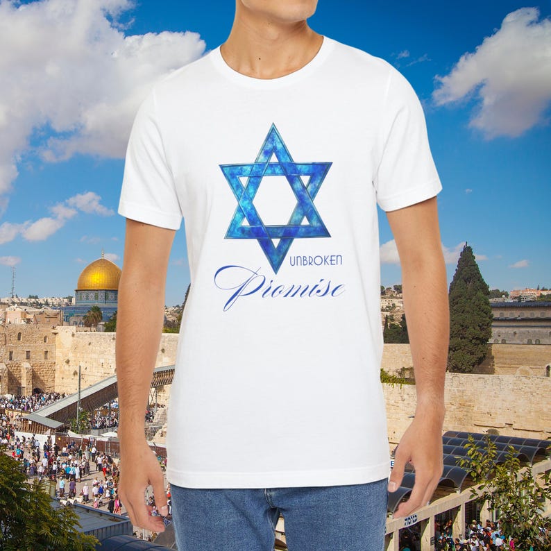 Pu&ograve; includere: T-shirt bianca con un disegno blu della Stella di David e la scritta "UNBROKEN Promise". La grafica &egrave; centrata sulla maglietta. Lo sfondo mostra un paesaggio urbano con una cupola dorata e una folla di persone.