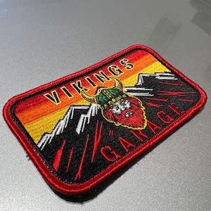 May include: Rectangular patch with a red border featuring the text "VIKINGS GARAGE". The patch has a Viking helmet design with a red beard, set against a mountain landscape with orange, yellow, and black colors.