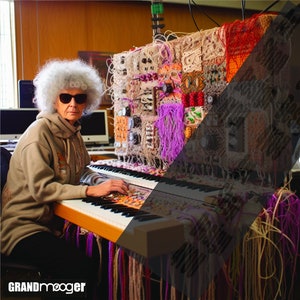 May include: A woman with gray hair and sunglasses plays a keyboard with a large, colorful, crocheted synthesizer behind it. The synthesizer is covered in yarn and fabric scraps. The text "GRANDmeager" is visible in the lower left corner of the image.
