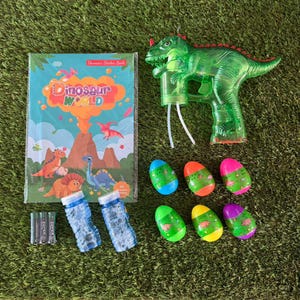May include: A children's dinosaur-themed playset featuring a green dinosaur-shaped bubble gun, a sticker book titled "Dinosaur World", six colorful egg-shaped containers, and two bubble wands. The items are arranged on a green surface.