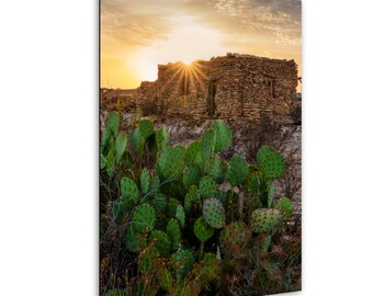 Puede incluir: Un edificio de piedra con un cactus en primer plano. El sol se pone detrás del edificio, proyectando un resplandor dorado en la escena.