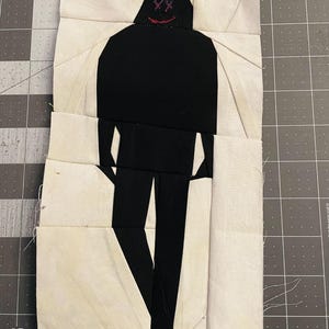 May include: A fabric art piece depicting a black figure with a hooded head and legs, set against a white and cream background. The figure's face has embroidered red details. The piece is constructed from various fabric pieces.