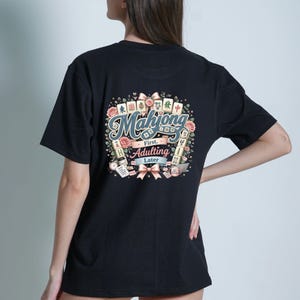 Op de afbeelding: Een zwart t-shirt met een afbeelding op de achterkant met de tekst Mahjong First, Adulting Later. Het ontwerp bevat illustraties van mahjongtegels, roze rozen, linten, een koffiemok, een laptop en een to-do lijst in een decoratieve opstelling.