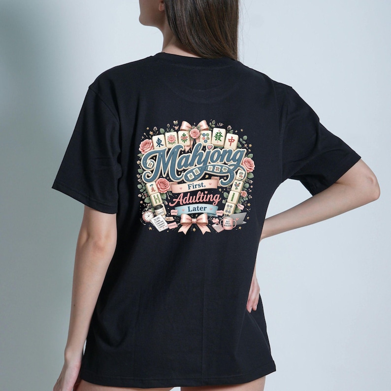 Op de afbeelding: Een zwart t-shirt met een afbeelding op de achterkant met de tekst Mahjong First, Adulting Later. Het ontwerp bevat illustraties van mahjongtegels, roze rozen, linten, een koffiemok, een laptop en een to-do lijst in een decoratieve opstelling.