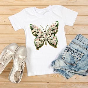 Op de afbeelding: Een wit T-shirt met korte mouwen en een grote afbeelding van een vlinder die is samengesteld uit verschillende mahjongstenen. De vlindervleugels zijn opgebouwd uit witte stenen met groene, rode en zwarte tekens en symbolen.