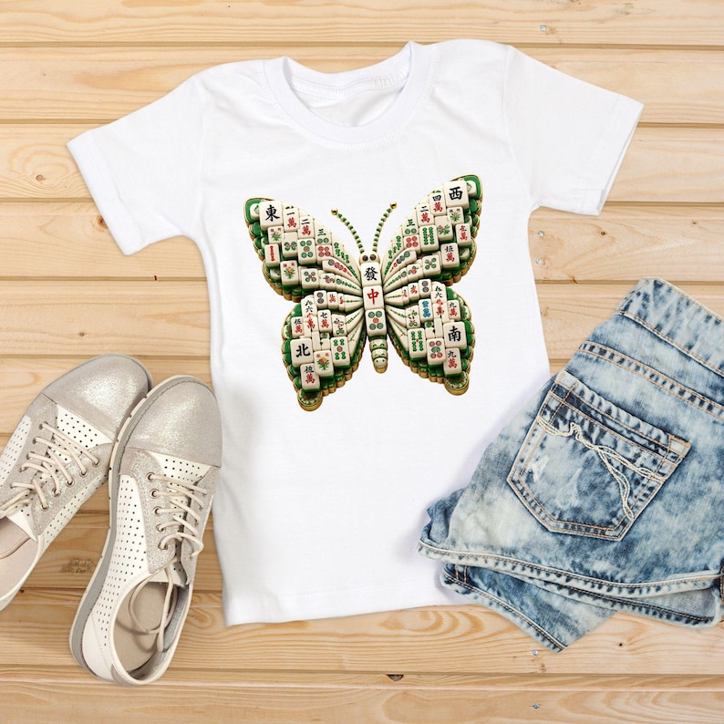 Op de afbeelding: Een wit T-shirt met korte mouwen en een grote afbeelding van een vlinder die is samengesteld uit verschillende mahjongstenen. De vlindervleugels zijn opgebouwd uit witte stenen met groene, rode en zwarte tekens en symbolen.