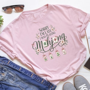Op de afbeelding: Een lichtroze t-shirt met een grafische print met de tekst "SORRY I'M LATE I WAS PLAYING Mahjong". Het ontwerp bevat illustraties van mahjongtegels, florale accenten en kleine roze strikjes die decoratief hangend zijn gerangschikt.