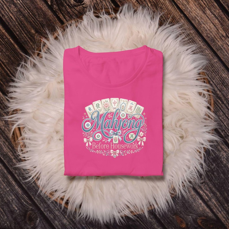 Op de afbeelding: Een felroze t-shirt met een grafische print van zes mahjongtegels in een boog boven de blauwe cursieve tekst Mahjong en de witte tekst Before Housework, omringd door decoratieve florale elementen en kleine strikjes.