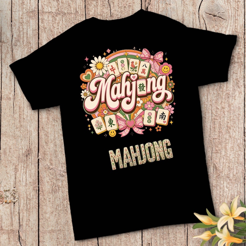 Op de afbeelding: Een zwart t-shirt met een kleurrijk grafisch ontwerp met het woord Mahjong in grote letters, omringd door illustraties van mahjongtegels, madeliefjes, hartjes, strikjes en smileys, met het woord MAHJONG in tegelletters eronder.