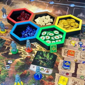 Könnte beinhalten: Ein Brettspiel mit verschiedenen Komponenten, darunter sechseckige Behälter mit roten Kristallen, beigen Steinen, blauen Edelsteinen, grünen Spielsteinen und Goldmünzen. Miniaturfiguren und Karten sind ebenfalls vorhanden, mit dem Titel "Min & Elwen".