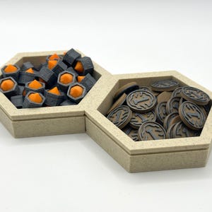 Puede incluir: Dos bandejas hexagonales beige llenas de piezas de juego. Una bandeja contiene piezas grises oscuras y naranjas, mientras que la otra contiene fichas circulares marrones con un diseño gris. Las bandejas están sobre una superficie blanca.