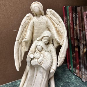 May include: A cream-colored angel figurine with outstretched wings, sheltering a smaller figure holding a baby. The sculpture has a carved appearance and is likely made of resin or a similar material. The figurine is a religious or decorative item.