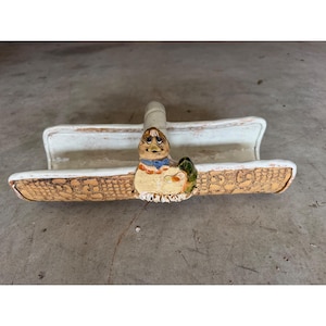 May include: A ceramic duck-shaped soap dish. The dish has a rectangular shape with a textured, basket-weave design on the sides. The duck has a blue scarf and a green wing. The dish is cream-colored with brown and yellow accents.