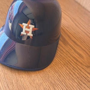 Op de afbeelding: Een marineblauwe honkbalhelm met het Houston Astros-logo aan de zijkant. Het logo heeft een witte "H" met een oranje ster. De helm staat op een houten oppervlak.