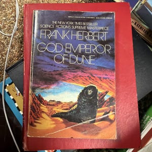 May include: A hardcover book titled "God Emperor of Dune" by Frank Herbert. The cover features a figure on a desert landscape with a purple and orange sky. The book is a New York Times bestseller and a science fiction masterpiece.